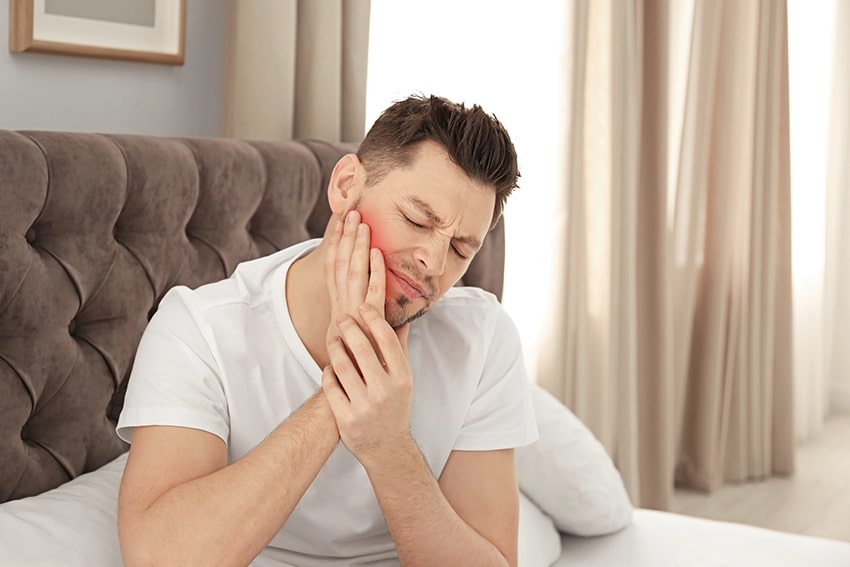Man holding jaw in pain while sitting on his bed. Jaw pain and TMJ can be treated at home.