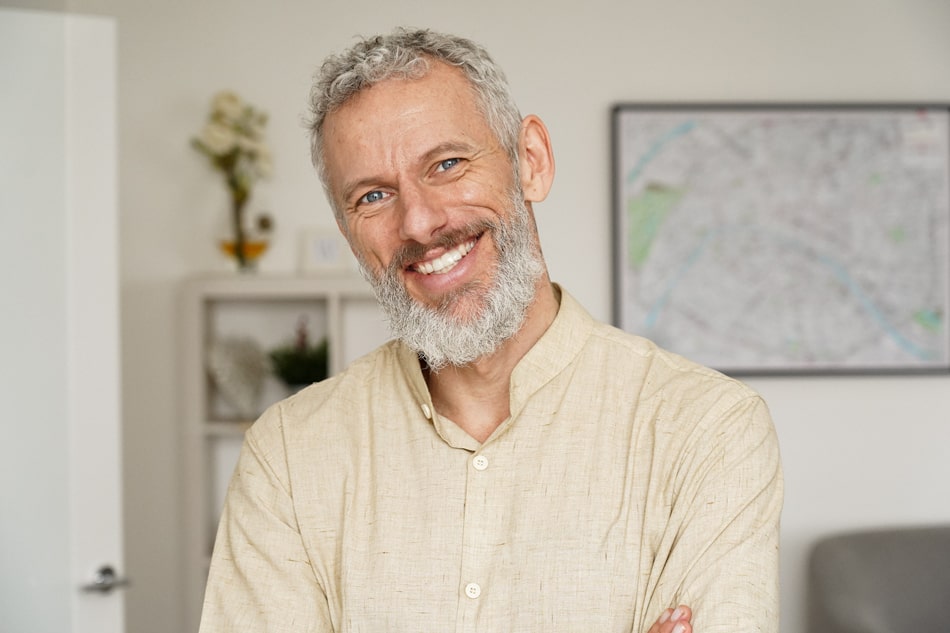An older man with a beard wearing an off-white button-up shirt smiling